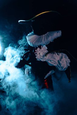 Yoğun mavi dumanın içinde duran gizemli soytarı karakterinin karanlık ve dramatik görüntüsü. Yoğun ışıklandırma, beyaz yaka ve uzun şapka. Cadılar Bayramı karakter teması. Yakın plan..