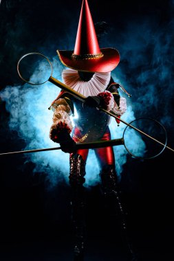 Mavi ışığa ve dumana karşı sirk aksesuarı tutan, özenle hazırlanmış kırmızı ve siyah kostümlü gizemli bir soytarının tüm vücudunu vuruyor. Cadılar Bayramı karakter teması.