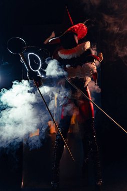 Sirk aksesuarı tutan kırmızı ve siyah kostümlü gizemli soytarı karakterinin dinamik çekimi. Yoğun beyaz duman, dramatik ışık ve yoğun ruh hali. Cadılar Bayramı karakter teması.