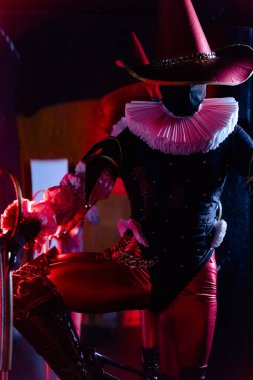 Kırmızı ve siyah kostümlü, uzun şapkalı ve beyaz yakalı esrarengiz bir soytarının kırmızı ışıklandırmalı dramatik çekimi. Cadılar Bayramı karakter teması. Yakın plan..