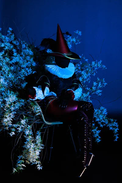 Siyah kostümlü gizemli soytarı karakter ve mavi ve beyaz ışıklı çiçeklerin arasında oturan uzun şapkalı. Dramatik ışıklandırma, karanlık fantezi havası. Cadılar Bayramı karakter teması.