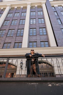 Siyah erotik iç çamaşırı ve deri trençkotlu şık ve cüsseli bir kadının son derece düşük açılı görüntüsü. Kendine güvenen kıvrımlı model kent binasının yanındaki korkuluklara dayanıyor, vücut pozitifliğini destekliyor.