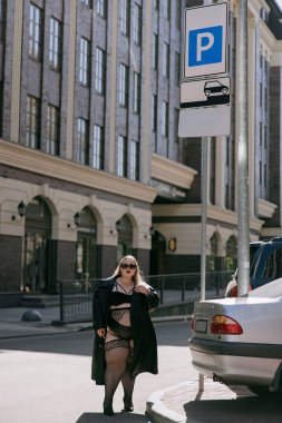 Siyahi erotik iç çamaşırlı ve deri trençkotlu şık ve kilolu bir kadının tam boy fotoğrafı. Kentteki park levhasının yanında poz veren kendine güvenen kıvrımlı model, vücut pozitifliğini ve modern tarzı destekliyor.