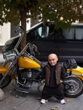 Cüce kılıklı gülen adam şehrin kaldırımında büyük sarı kruvazör motosikletinin yanında duruyor. Günlük kıyafetler, kendine güvenen pozlar, benzersiz bireyler ve ulaşım..
