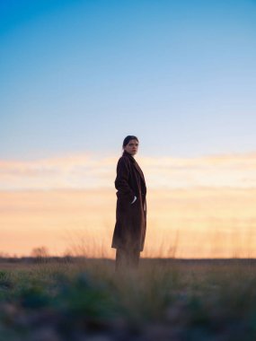 Alçak açı tam boy portre. Soğuk havada, parlak altın saat günbatımında ya da gün doğumunda gökyüzü arka planında ayakta duran genç kadın profili silueti..