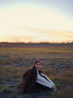 Çayırda çimenlerde oturan genç bir kadın altın saat gün batımında sıcak ceket ve kazak giyiyor ya da gün doğumunda kameraya bakıyor..