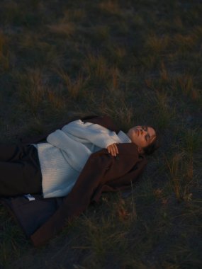 Gün batımında gözleri kapalı çim tarlasında dinlenen genç bir kadının huzurlu görüntüsü. Model beyaz süveter ve koyu renk ceket giyer, doğada huzur, rahatlık ve sükuneti ifade eder.