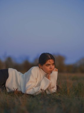 Sonbahar alacakaranlığı sırasında çim tarlasında rahat beyaz bir kazak giyen düşünceli genç bir kadın. Başını çevirir, hayatı düşünür, ciddi ve huzurlu bir ruh hali içindedir..
