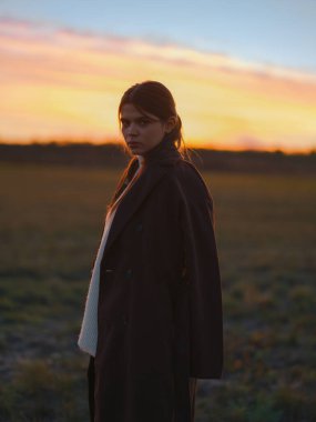 Ciddi genç kadın dramatik turuncu ve pembe günbatımı gökyüzüne karşı sahada duruyor. Beyaz süveterin üzerine koyu renk bir ceket giyer, kameraya özgüvenle bakar ve kendini ifade eder..