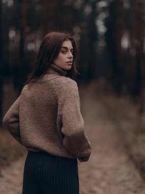 Genç bir kadın orman yolunda dikiliyor ve arkasını kolluyor. Moda, tarz ve doğal sonbahar havası..