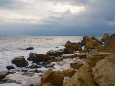 Rocky Karadeniz kıyıları dramatik bir gökyüzü altında. Kıyı boyunca çok sayıda büyük ve küçük kaya parçacıkları, güçlü dalgaların oluşturduğu yoğun deniz köpüğüyle kaplıydı. Vahşi doğa, erozyon, soğuk mevsim.