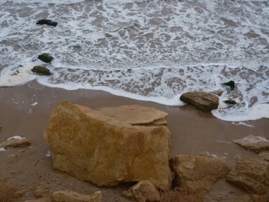 Islak kumlu bir sahildeki büyük sarı kumtaşı kayanın yukarıdan görünen görüntüsü. Beyaz deniz köpüğü ve su güçlü dalgalardan çekiliyor. Kıyı doğası, soyut doku, soğuk mevsim.