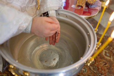 Papazın ellerinin suyun üzerinde küçük gümüş haç kolyesini büyük vaftiz fontuyla mumlarla tutarken yakın plan. Kutsal ayine, haça ve vaftiz törenine odaklan..