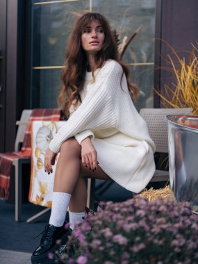 Beyaz örgü süveter ve platform ayakkabılı şık bir kadın açık kafede oturuyor, sonbahar çiçekleri ve mevsimlik dekorasyonlarla çevrili..