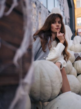 Güzel, gri ceketli ve beyaz kazaklı genç bir kadın beyaz balkabaklarının arasında parmaklarıyla poz vererek büyüleyici Cadılar Bayramı atmosferi yaratıyor..