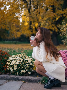 Parkta çömelmiş gülümseyen genç bir kadın, elinde kahve fincanı, renkli sonbahar yapraklarının ve çiçek açan kasımpatıların tadını çıkarıyor, rahat beyaz örgü elbiseler giyiyor..