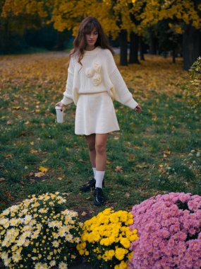 Beyaz örgü elbiseli ve süveterli güzel bir kadın. Elinde çiçeklerle parkta yürüyor. Elinde kahve, renkli sonbahar yaprakları ve çiçek açan annelerle çevrili..