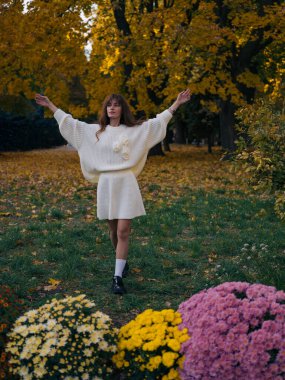 Parkta kolları havada duran güzel genç bir kadın sonbahar yapraklarının, sarı ağaçların ve çiçeklerin tadını çıkarıyor. Sonbaharın özgürlüğünü ve güzelliğini kutluyor..