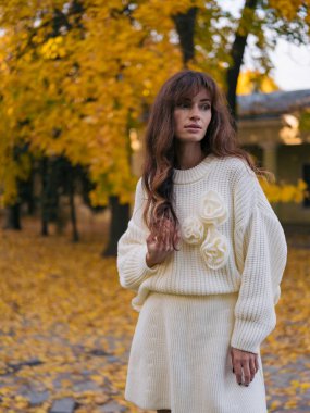 Beyaz örgü süveter ve etek giymiş güzel bir kadının portresi. Parkta duruyor, gözlerini kaçırıyor. Etrafı parlak sarı sonbahar yeşillikleriyle çevrili..