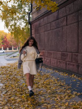 Beyaz örgü takım elbiseli şık genç bir kadın ve gri ceketli. Sarı yapraklarla kaplı kaldırımda neşeyle yürüyor. Elinde kahve fincanı ve siyah bir çantayla..