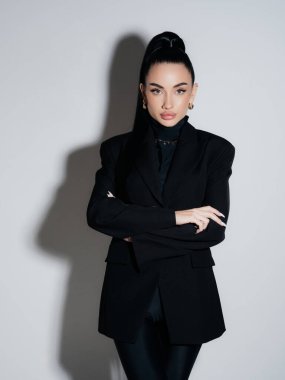 Siyah ceketli balıkçı yaka giyen zarif genç bir kadın çapraz kollarla poz veriyor. Kendine güvenen ciddi yönetici portresi stüdyosu çekimi. Güç stili moda güzellik konsepti.