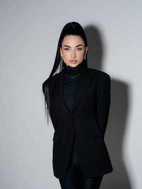 Siyah ceketli ve balıkçı yakalı zarif genç bir kadın elleri arkada duruyor. Kendine güvenen ciddi kadın yönetici profesyonel model stüdyo çekimi. Güç stili moda güzellik konsepti.
