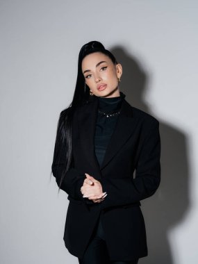 Siyah ceketli ve boğazlı kazaklı zarif genç bir kadın elleri bağlı duruyor. Kendine güvenen ciddi kadın yönetici profesyonel model stüdyo çekimi. Güç stili moda güzellik konsepti.