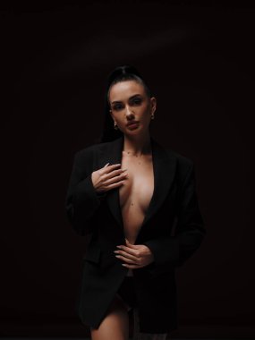 Siyah düğmesiz ceketli, koyu arkaplanda poz veren şık ve kendine güvenen çıplak bir kadın. Kadın gücü, cesur moda ve ticari marka konsepti..