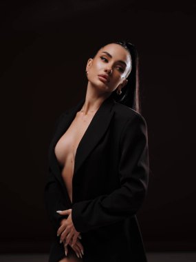 Siyah düğmesiz ceketli, koyu arkaplanda poz veren şık ve kendine güvenen çıplak bir kadın. Kadın gücü, cesur moda ve ticari marka konsepti..