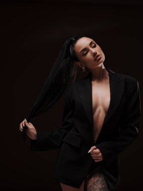 Siyah düğmesiz ceketli, koyu arkaplanda poz veren şık ve kendine güvenen çıplak bir kadın. Kadın gücü, cesur moda ve ticari marka konsepti..