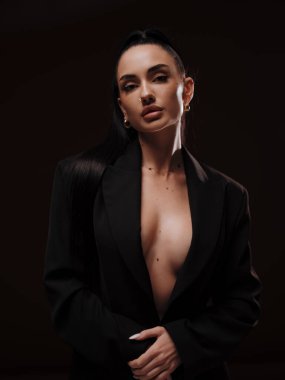 Siyah düğmesiz ceketli, koyu arkaplanda poz veren şık ve kendine güvenen çıplak bir kadın. Kadın gücü, cesur moda ve ticari marka konsepti..