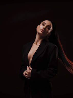 Siyah düğmesiz ceketli, koyu arkaplanda poz veren şık ve kendine güvenen çıplak bir kadın. Kadın gücü, cesur moda ve ticari marka konsepti..