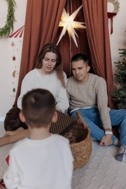Genç anne ve baba, terracotta gölgesinin altında birlikte oturmuş hediye sepetini keşfediyorlar. Küçük oğulları ön planda odakta değil. Şenlikli dekorasyonları olan rahat İskandinav evi..