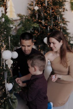 Küçük oğullarının Noel ağacına beyaz süsler asmasını izlerken gülümseyen genç ebeveynler. Oda şenlikli dekorasyonlar, sıcak ışıklar ve mücevherlerle dolu, rahat bir tatil ortamı yaratıyor..