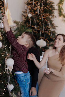 Küçük çocuk uzun Noel ağacına süs asmak için uzanıyor. Annem yanında oturuyor, gülümsüyor ve daha çok mücevher taşıyor. Ağaç, beyaz ve altın süsler ve parlayan ışıklarla süslenir..