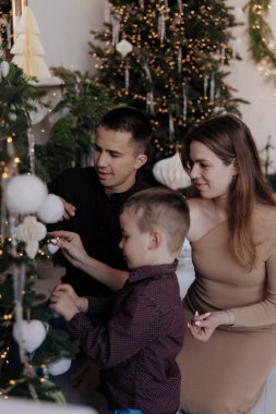 Genç ebeveynler küçük oğullarının Noel ağacına beyaz ve altın süsler asmasına yardım ediyorlar. Sahne ılık ışıklar, şenlikli mücevherler ve parlak İskandinav iç kısımlarında sıcak tatil atmosferiyle dolu.