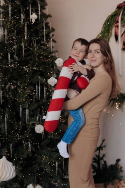 Küçük oğlunu kollarında tutan gülümseyen bir anne. Çocuğun elinde büyük, pelüş çizgili şeker kamışı var. Noel ağacının yanında süslerle, mücevherlerle ve ışıklarla süslenmiş bir odada duruyorlar..