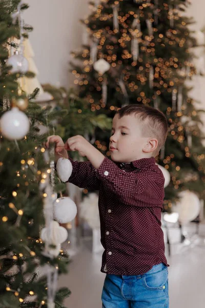 Küçük çocuk Noel ağacına beyaz süsler ve takılar asıyor. Arka plan sıcak altın ışıklarla ve rahat bir evde şenlik havasıyla dolu..