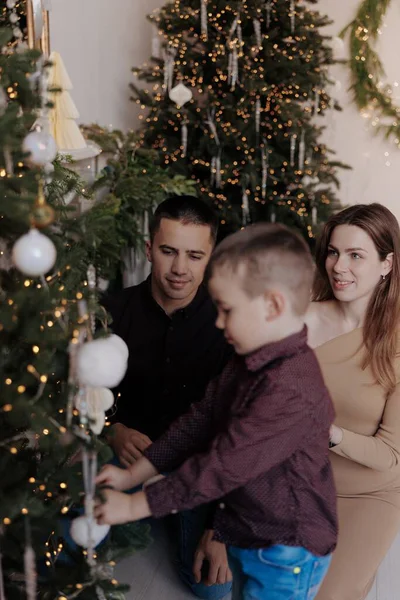 Küçük oğullarının Noel ağacına beyaz süsler asmasını izleyen genç ebeveynler. Sıcak ışıklarla, şenlikli mücevherlerle ve İskandinav iç mekanlarıyla sıcacık bir tatil ortamı..