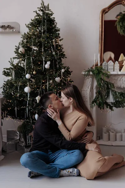 A young man and woman in a romantic embrace, kissing while sitting on the floor in a festive room. Beside them is a tall Christmas tree decorated with white ornaments, baubles, and glowing lights.