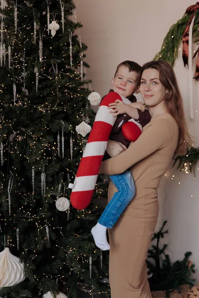 Küçük oğlunu kollarında tutan gülümseyen bir anne. Çocuğun elinde büyük, pelüş çizgili şeker kamışı var. Noel ağacının yanında süslerle, mücevherlerle ve ışıklarla süslenmiş bir odada duruyorlar..