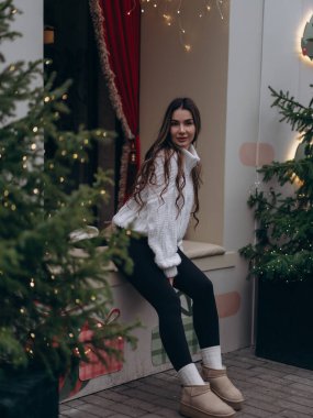 Süslü süveter ve siyah tayt giyen mutlu genç kadın dışarıda, dekore edilmiş pencerenin yanında oturuyor. Noel ağaçları, Bokeh ışıkları ve kırmızı perdeleri olan şenlikli kış atmosferi.