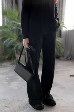Kadın elinin şık, siyah deri bir ekmek torbası tutarken çekilmiş ayrıntılı bir fotoğrafı. Siyah balıkçı yaka ve geniş paçalı pantolon giyen bir model. Minimalist moda aksesuarları ve şehir tarzı konsept parlak bir iç mekan..