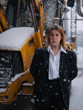 Kar yağışı sırasında dışarıda siyah takım elbiseyle poz veren şarap lekeli bir iş kadını. Sarı endüstriyel makineler arka planında, kentsel ortamda portre..