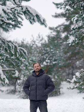 Donmuş ormanlık alanda poz veren gri balon giyen mutlu genç adam. Yakışıklı bir adam ve karla kaplı ağaçlarla kış mevsimi açık hava yaşam tarzı. Soğuk havadan hoşlanan birinin portresi..