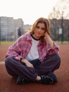 Pembe ekose gömlekli güzel kız dışarıda basketbol sahasında bacak bacak üstüne atmış poz veriyor. Gün batımında kırmızı kauçuk kırıntıları üzerinde oturan kadın model. Şehir yaşam tarzı ve moda.