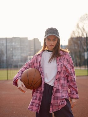 Pembe ekose gömlekli ve şapkalı çekici bir kadın manken basketbolla ayakta duruyor. Gün batımında dışarıda poz veren güzel bir kız. Sportif şehir yaşam tarzı, gençlik modası ve hobi konsepti.