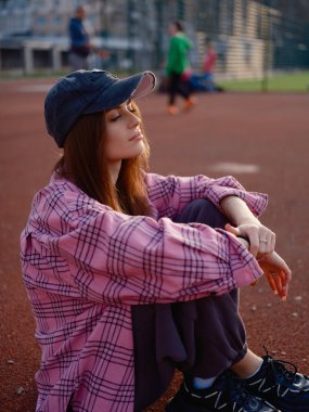 Pembe ekose gömlekli ve şapkalı, spor sahasında oturan barışçıl kadın model. Gün batımında kapalı gözlerle gün ışığının tadını çıkaran güzel bir kız. Kentsel yaşam tarzı, dinginlik ve moda konsepti.