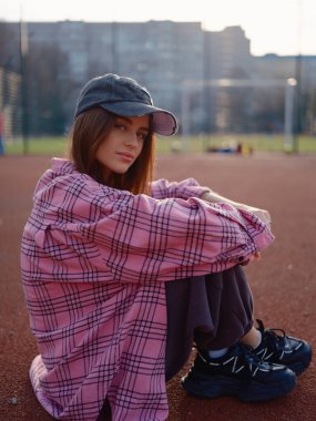 Pembe ekose gömlekli ve şapkalı, spor sahasında oturan barışçıl kadın model. Gün batımında kameraya bakan güzel bir kız. Kentsel yaşam tarzı, dinginlik ve moda konsepti.