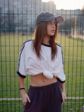 Güneş batarken tel örgülerin yanında duran beyaz tişörtlü ve beyzbol şapkalı güzel bir kız. Açık hava futbol sahasında ince beli olan bir kadın model. Şehir modası.